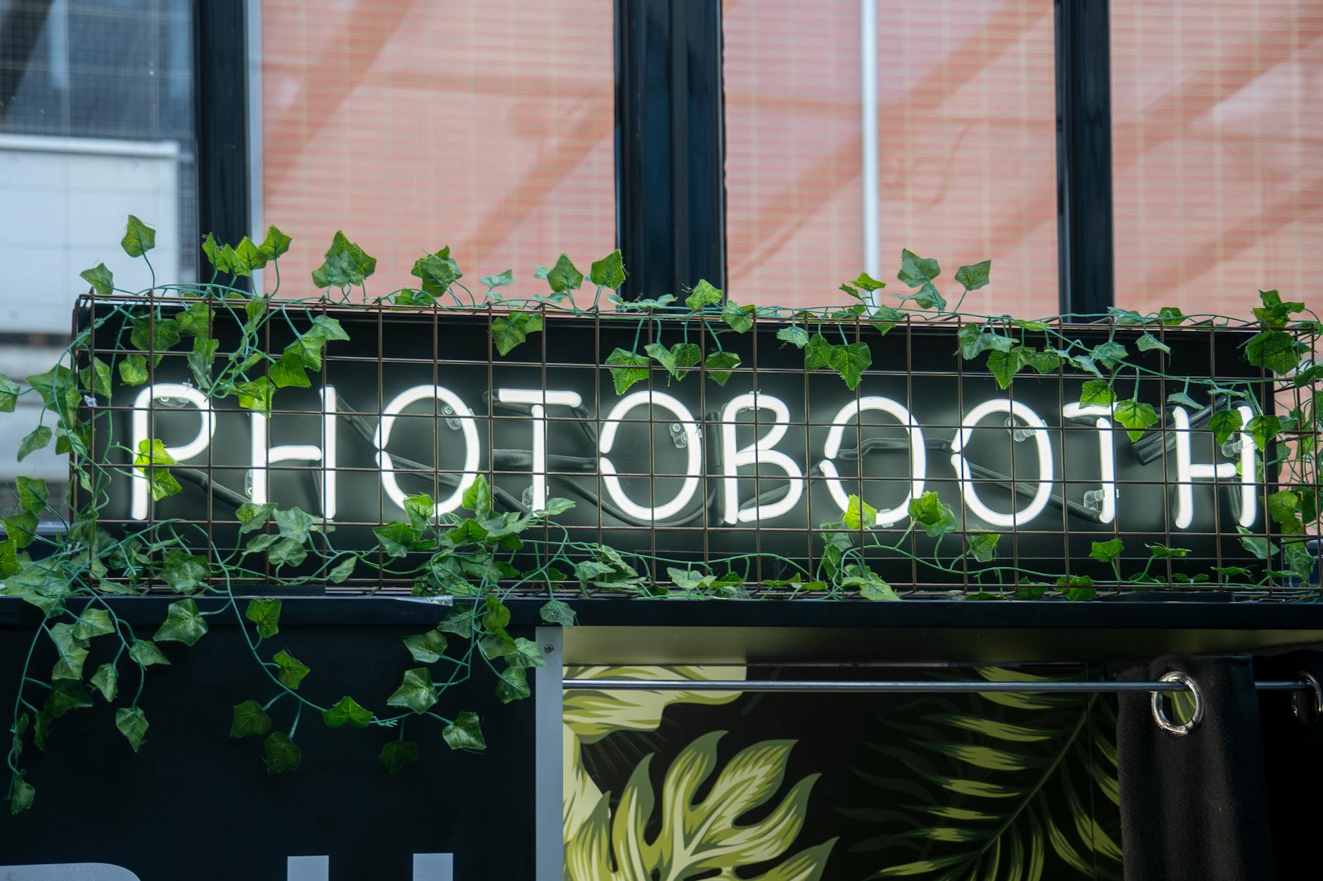 A photobooth sign with neon lights and greenery in an urban environment. Photobooth festival