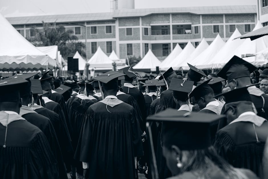 Diverse group of graduates in gowns at an outdoor ceremony with tents and buildings.