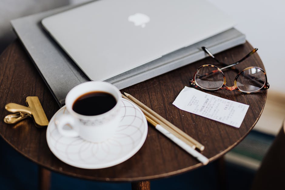 Gestion administrative et comptable A close-up of a wooden table with a laptop, coffee, eyeglasses, and stationery, creating a cozy workspace vibe.